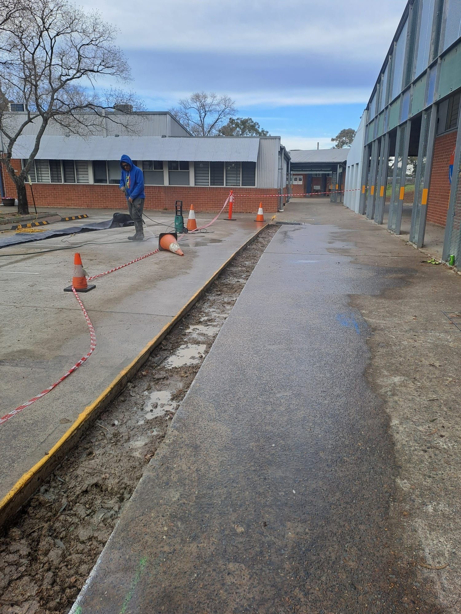 Concrete path works with a worker pressure cleaning behind red and white barrier tape and traffic cones