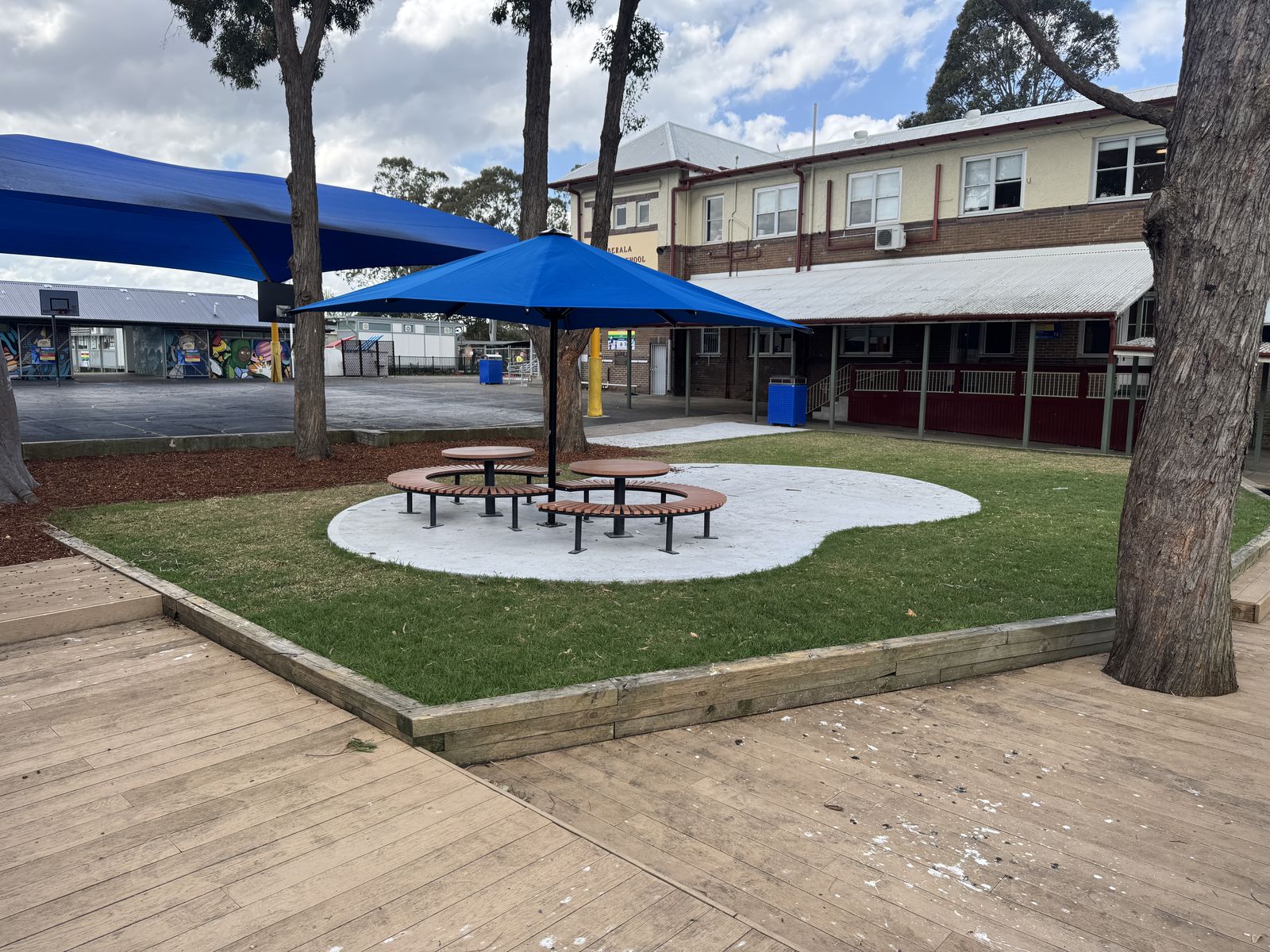Timber decking beside new turf and outdoor bench seating under blue shade umbrellas in a courtyard