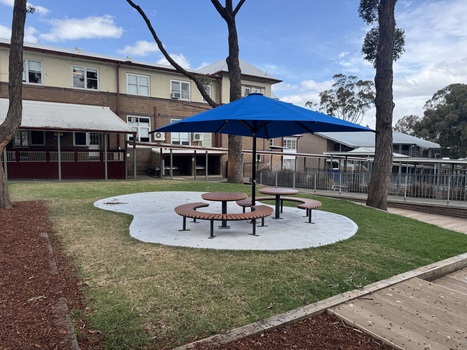 Curved timber bench seating and table under a blue umbrella on a concrete pad, with new turf and timber decking
