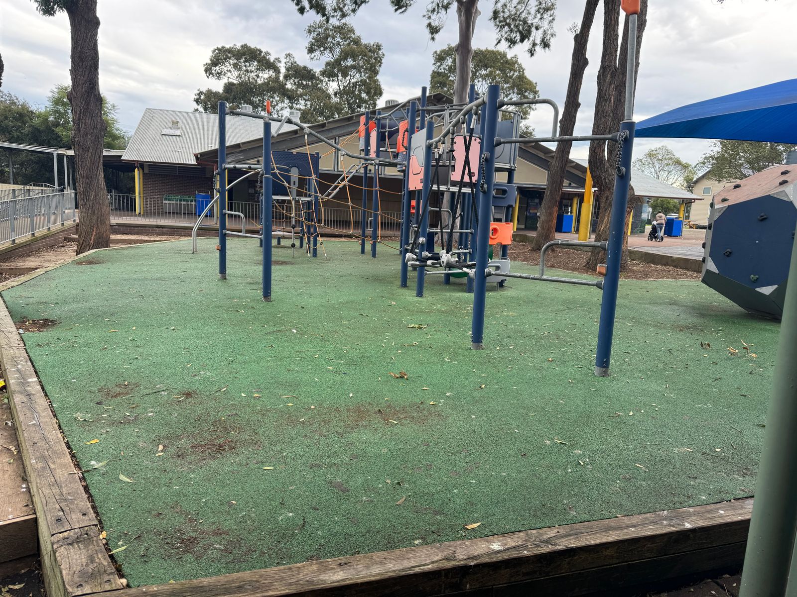 Playground equipment on a green softfall surface with a blue shade structure behind